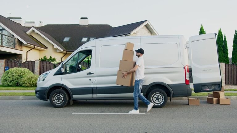 Hombre cargando cajas de cartón hacia una furgoneta blanca grande con las puertas traseras abiertas