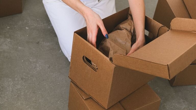 Manos colocando objetos envueltos en papel kraft dentro de una caja de cartón de mudanza