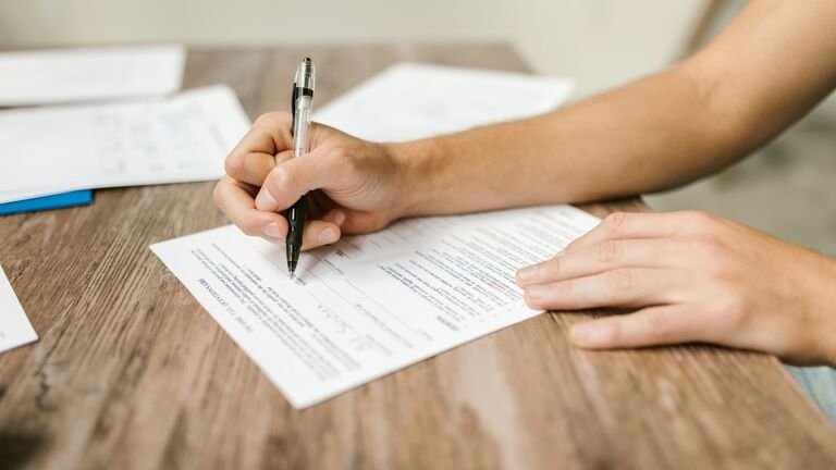 Mano rellenando con bolígrafo un formulario en papel sobre una mesa de madera, junto a otros documentos