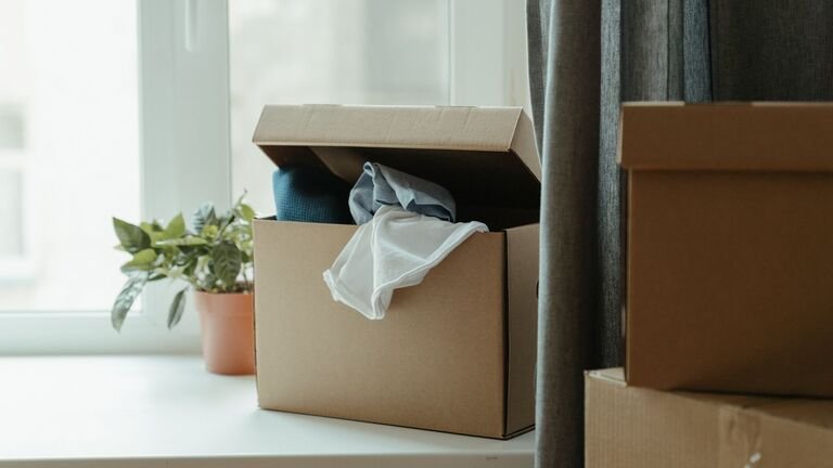 Caja de mudanza abierta con ropa junto a una ventana, al lado de otras cajas de cartón cerradas