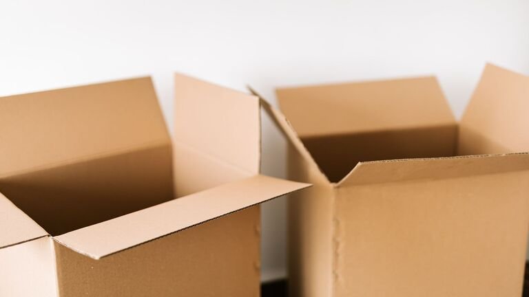 Dos cajas de cartón abiertas y vacías sobre una superficie oscura, delante de una pared blanca